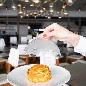 A hand lifting a cloche off a plate of pasta in a modern restaurant with white chairs and hanging lights in the background.
