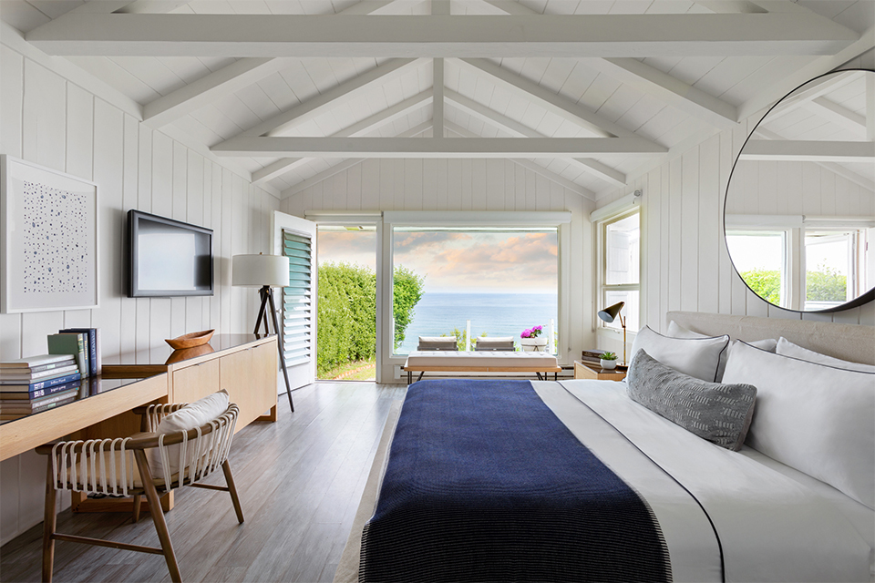 Cozy, modern bedroom with a large bed, blue blanket, and ocean view through open glass doors. Decor includes a desk, TV, and round mirror.