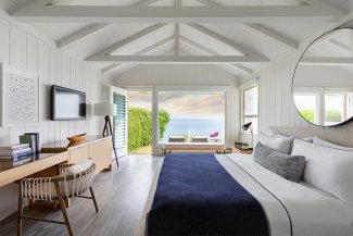 Cozy, modern bedroom with a large bed, blue blanket, and ocean view through open glass doors. Decor includes a desk, TV, and round mirror.