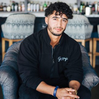 Young man with a curly hairstyle sitting on a gray chair, wearing a black jacket with "Savage" embroidery, in a relaxed indoor setting with a bar background.