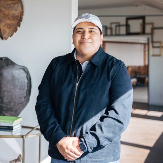 Smiling person in a blue jacket and cap stands indoors near a table with books, in a room with framed pictures on the wall.