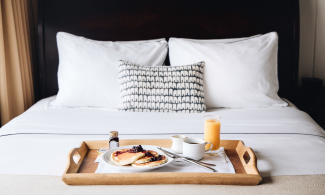 Breakfast tray with pancakes, juice, and coffee on a neatly made bed with white pillows and a patterned cushion.